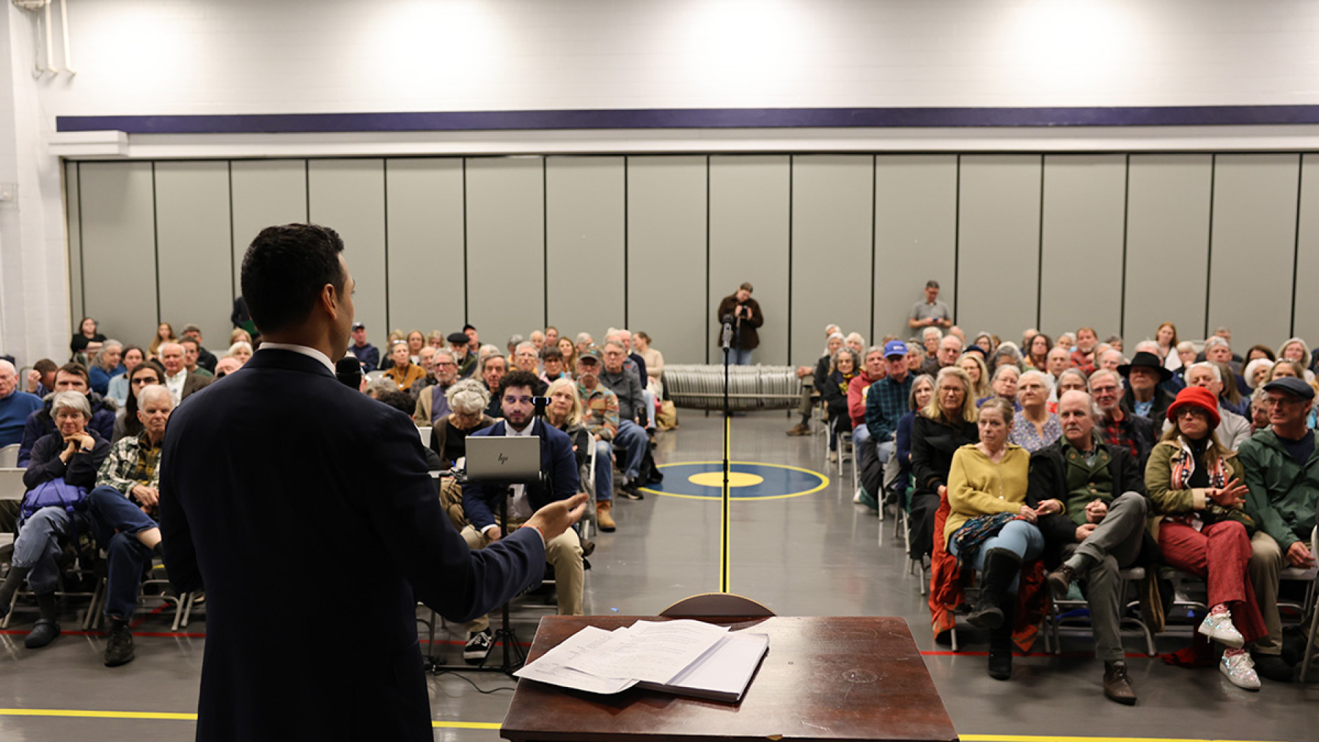 Congressman Subramanyam stands at a podium in a gym-style hall, holding a microphone as he addresses a large seated audience