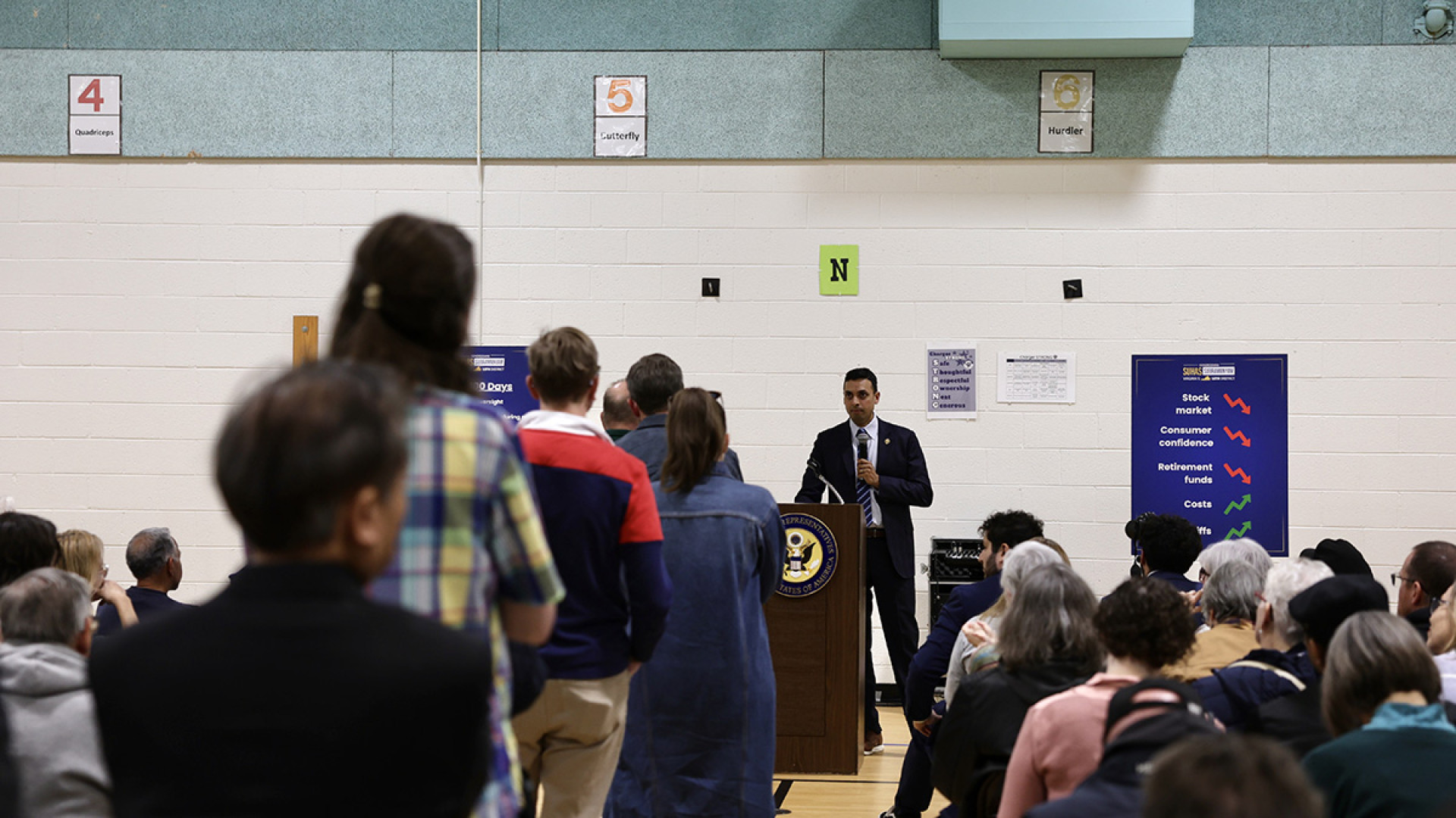 Congressman Suhas Subramanyam stands at a podium in a school gym, microphone in hand, as constituents line up in front of him to ask questions.