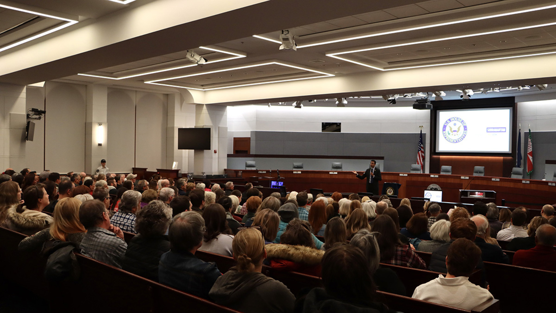 Congressman Suhas Subramanyam addresses a full chamber-style audience from the front of a formal hearing room, gesturing as he speaks.