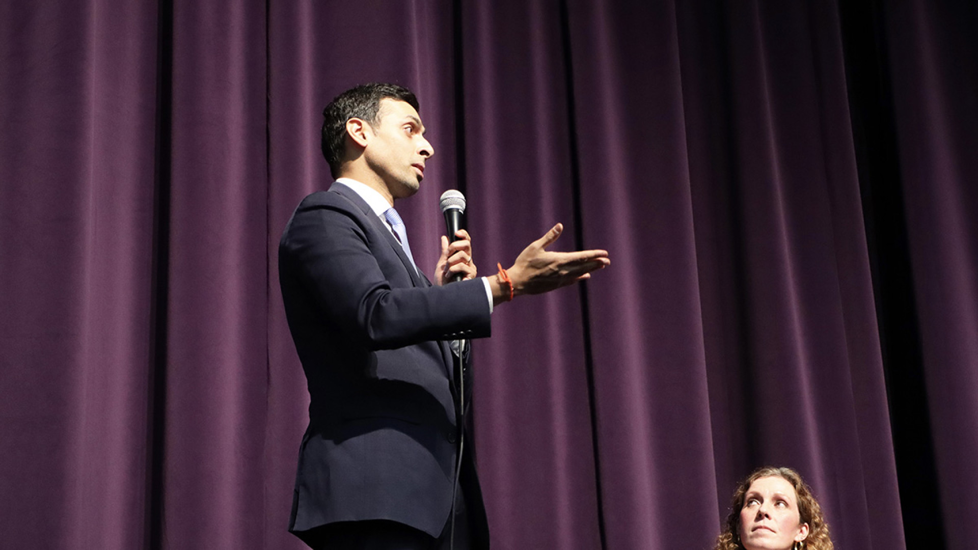 Congressman Suhas Subramanyam gestures thoughtfully into a handheld microphone as he speaks on stage before a purple curtain, while a seated panelist looks on.