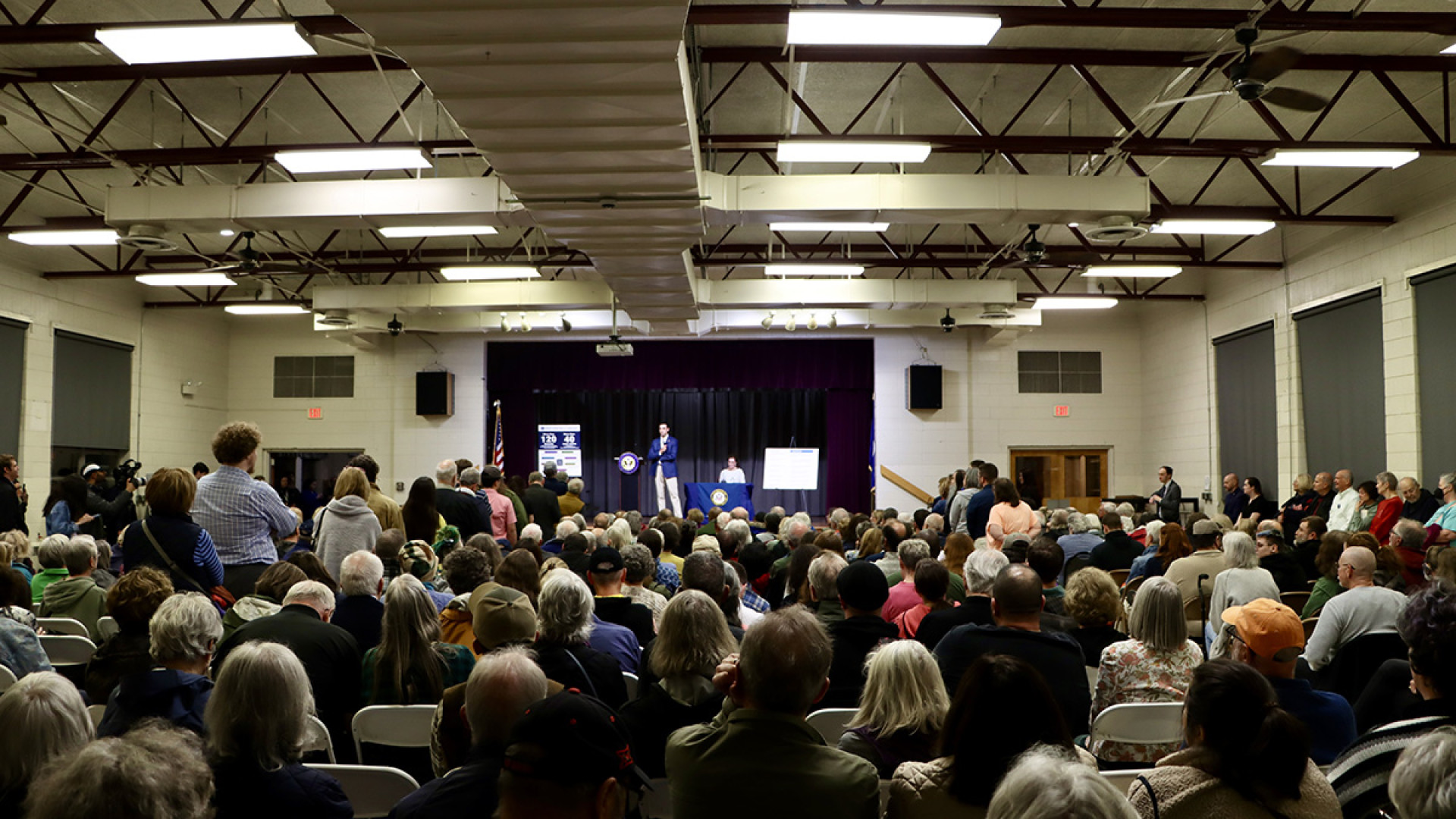 Congressman Suhas Subramanyam stands on a stage in a crowded community hall, addressing a packed audience of seated and standing constituents.