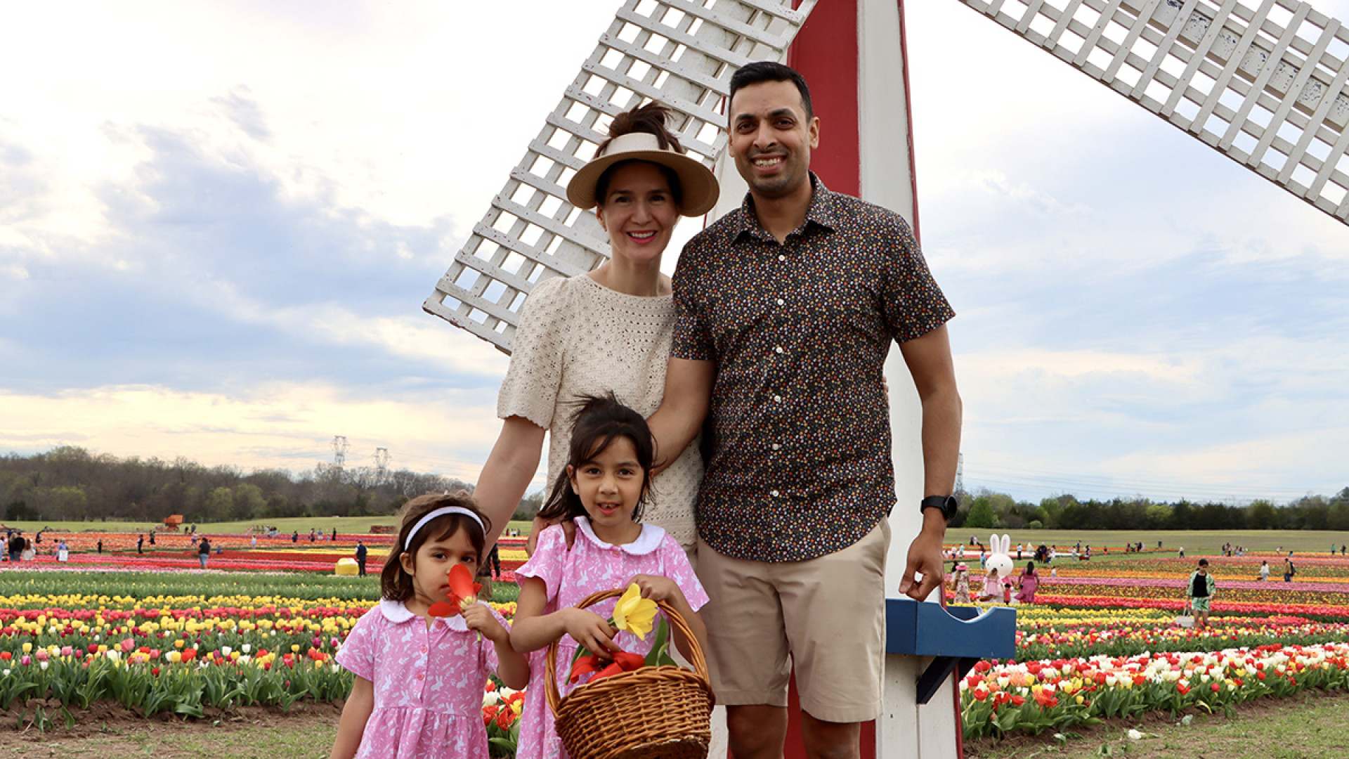 Congressman Suhas Subramanyam and his wife stand behind their two young daughters—each holding a basket of tulips—in front of a white-and-red windmill set against rows of colorful blooms.